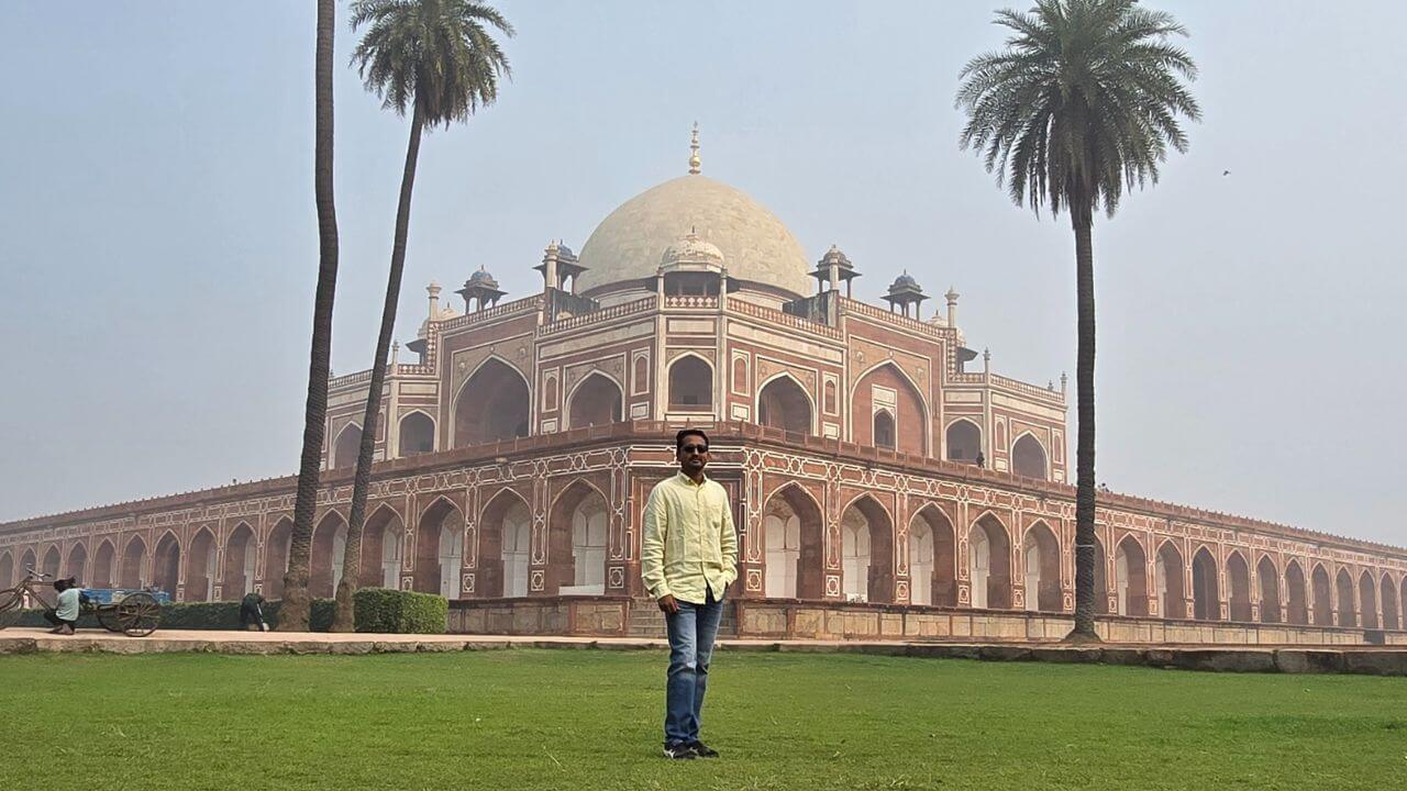 A visitor stands on the lawn facing Humayun’s Tomb in Delhi, with tall palm trees on both sides, captured on a clear day.