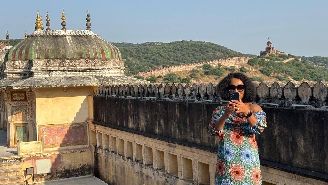A woman taking a photo on the terrace of Amber Fort in Jaipur with surrounding hills and historic fort walls in the background.
